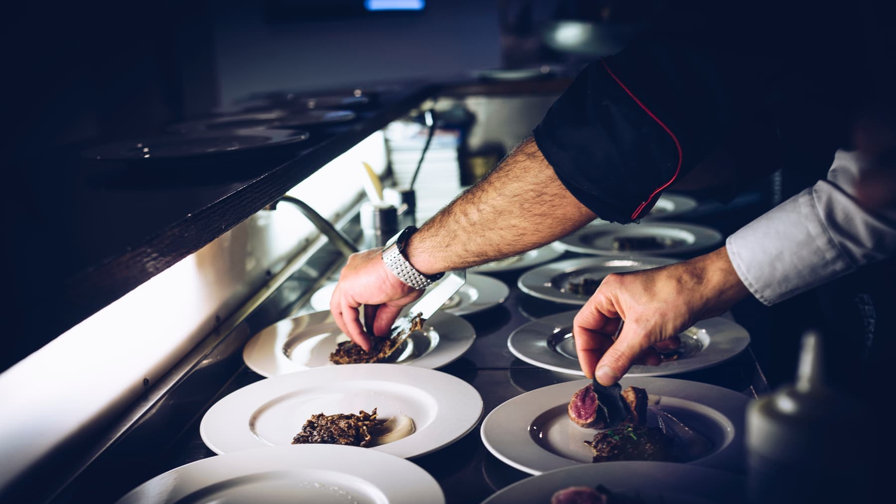Hender som anretter kjøtt og tilbehør på rader med hvite tallerkener ved passet i et restaurantkjøkken.