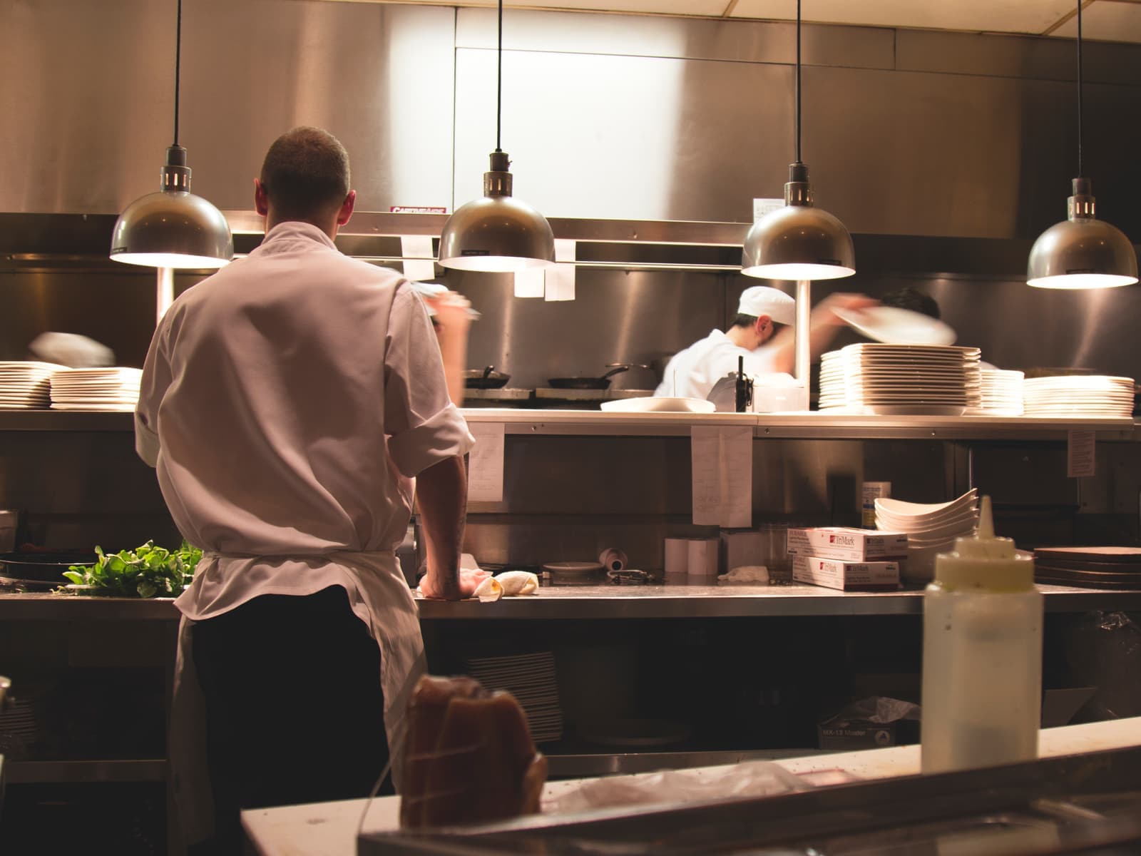 Kokk i hvit jakke sett bakfra ved passet i et travelt restaurantkjøkken, med varmelamper og stabler med tallerkener.