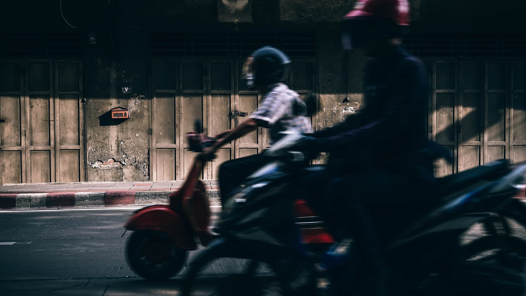 To førere med hjelm suser forbi på scooter og motorsykkel i en skyggefull bygate, uskarpe i fart.