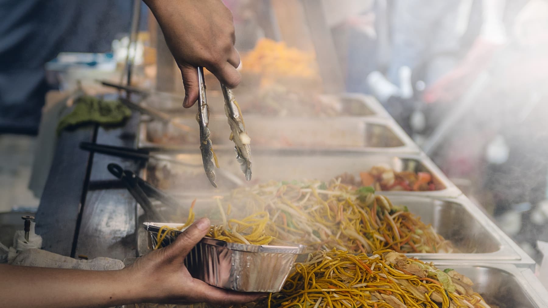 Stekte nudler øses med tang over i en takeaway-form av aluminium ved en travel matbod.