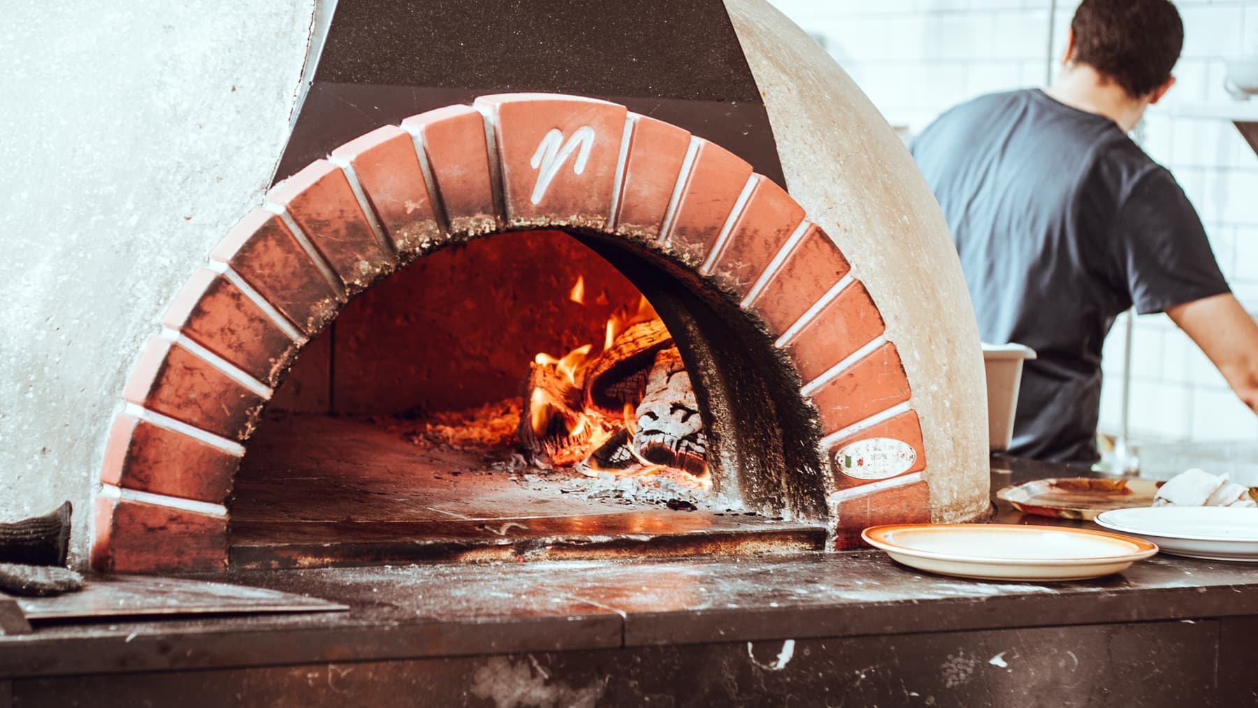 Vedfyrt pizzaovn i murstein med flammer innerst, mens en kokk jobber ved benken i bakgrunnen.